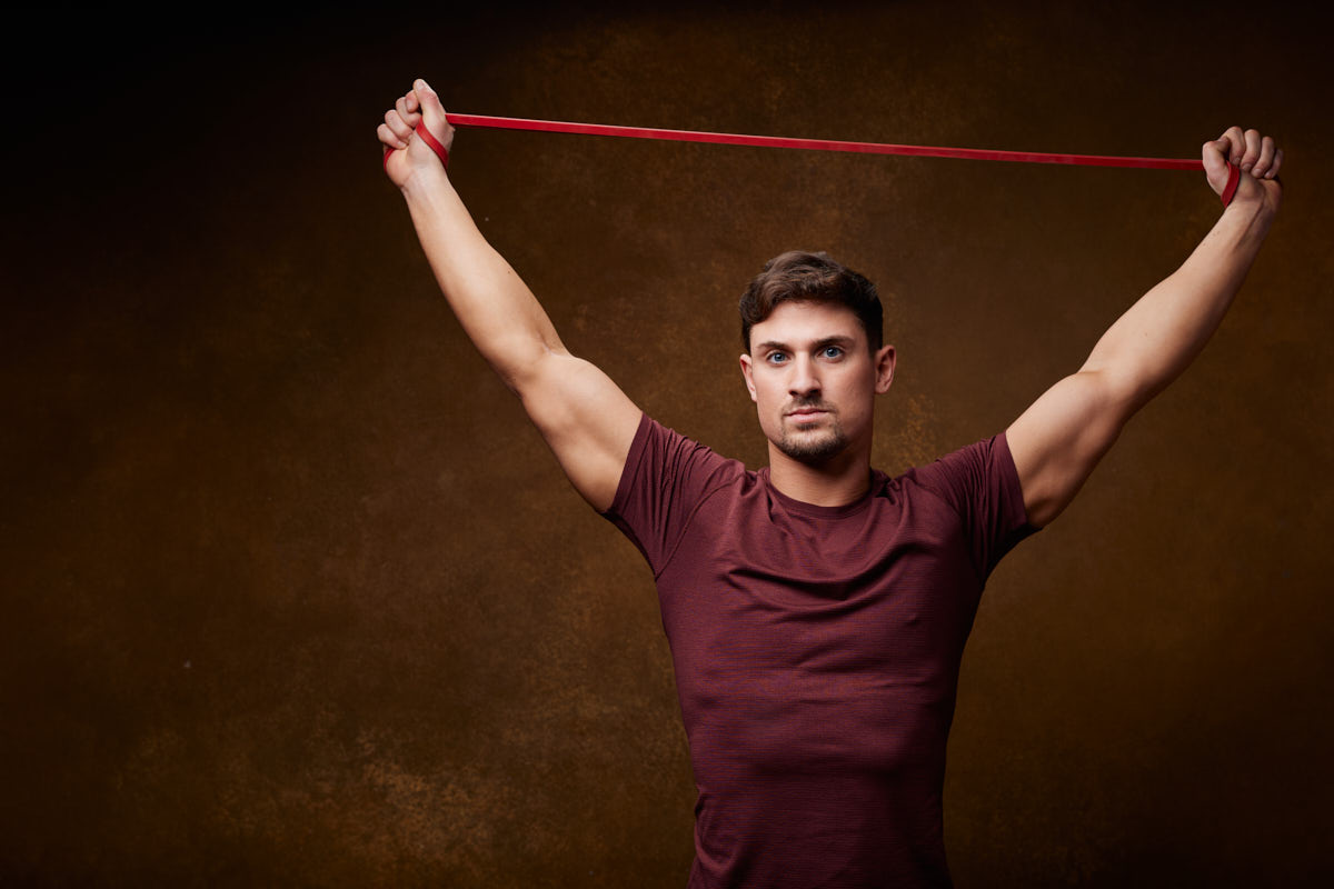 In this image, a fitness model in a maroon shirt and gray shorts is holding a red resistance band above his head with both hands, appearing to stretch or exercise.
