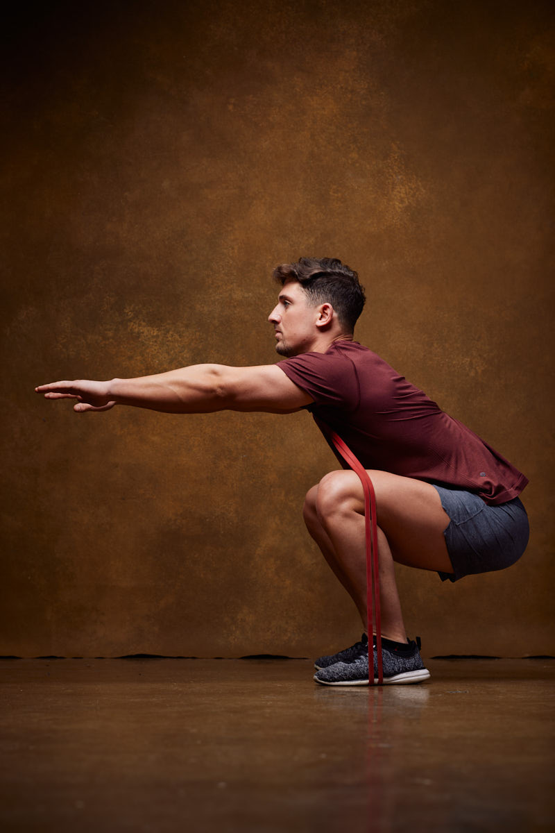 This image shows a model in a maroon shirt and gray shorts performing a deep squat with his arms extended forward. He is using a red resistance band around his legs to add difficulty to the exercise.