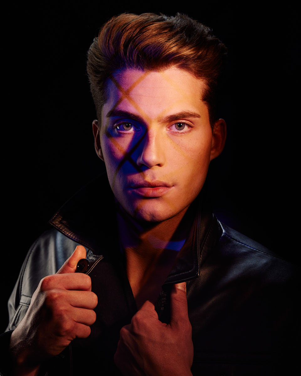 Christian is wearing a black leather jacket with the collar up, giving a confident and edgy look. The image features artistic lighting with shadows crossing his face, adding a dramatic and stylish effect.