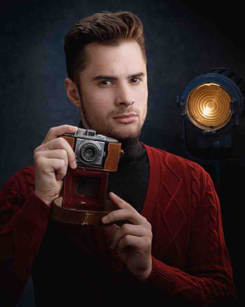 A close-up portrait of Saxxon holding a vintage camera, wearing a red sweater. The background includes a professional studio light, adding a dramatic effect to the portrait.