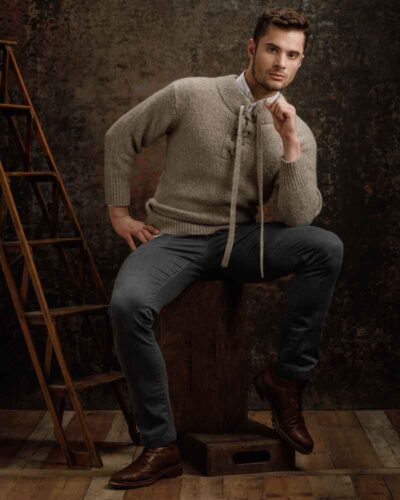 Saxxon is sitting on a wooden box, dressed in a light gray sweater and dark gray pants. The background features a rustic, dark textured backdrop, complementing his casual yet sophisticated look.