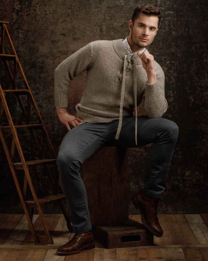 Saxxon is sitting on a wooden box, dressed in a light gray sweater and dark gray pants. The background features a rustic, dark textured backdrop, complementing his casual yet sophisticated look.