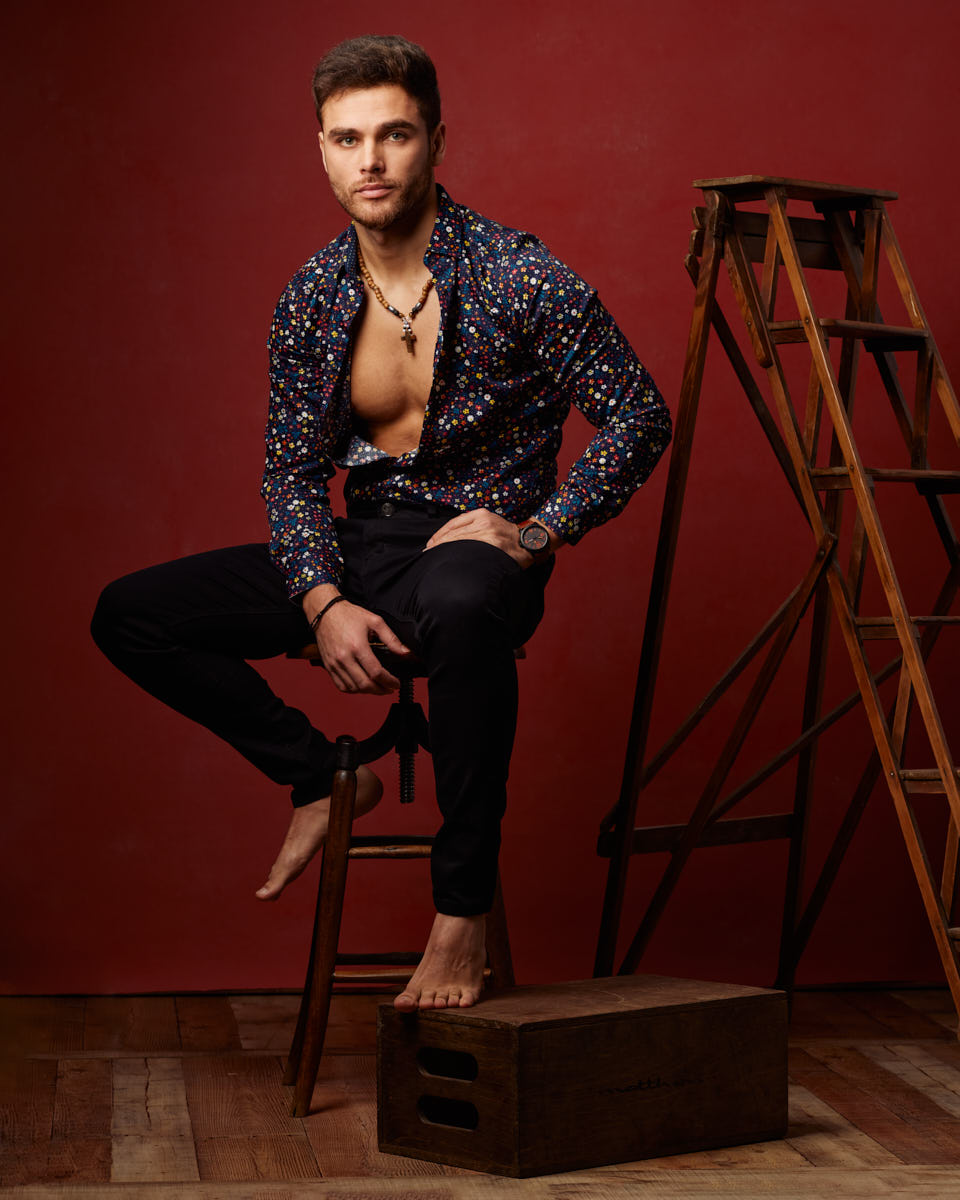 This is a full body portrait of Constantine in dark jeans, and a printed long sleeve shirt, which is open, exposing his chest. He is seated on an antique wooden store in front of an old wooden ladder in front of a red painted canvas backdrop showcasing Studio model photography in Chicago