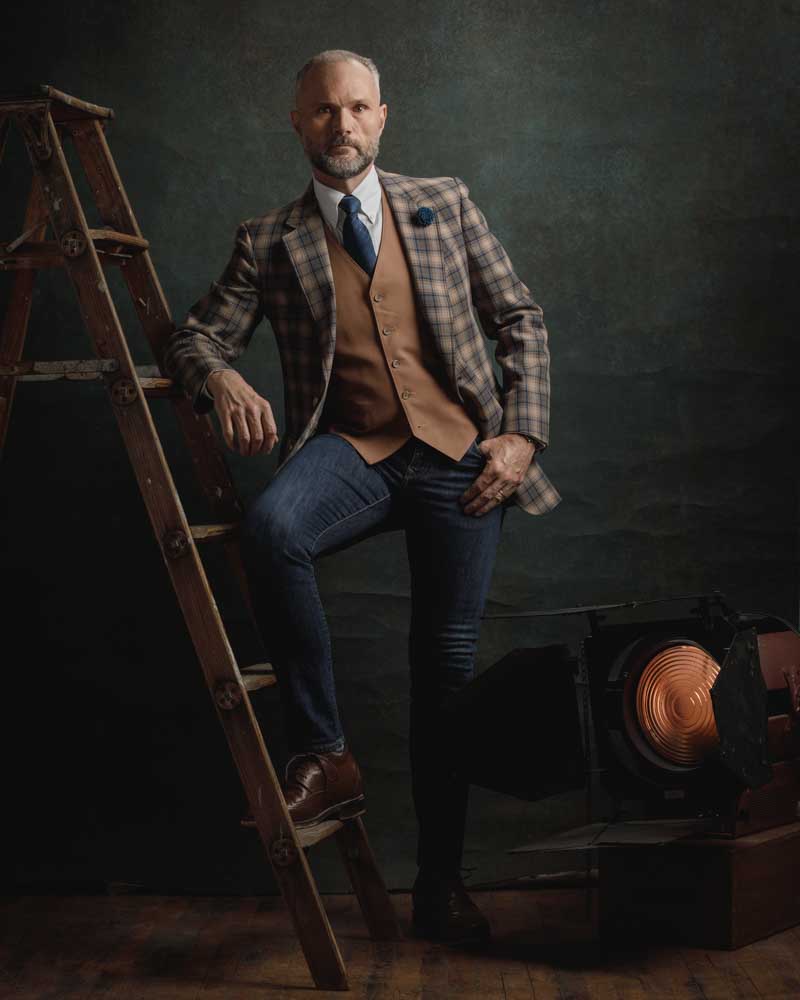 Wayne is leaning against a wooden ladder, dressed in a plaid jacket and a vest, with a vintage spotlight adding to the rustic atmosphere of the shot.