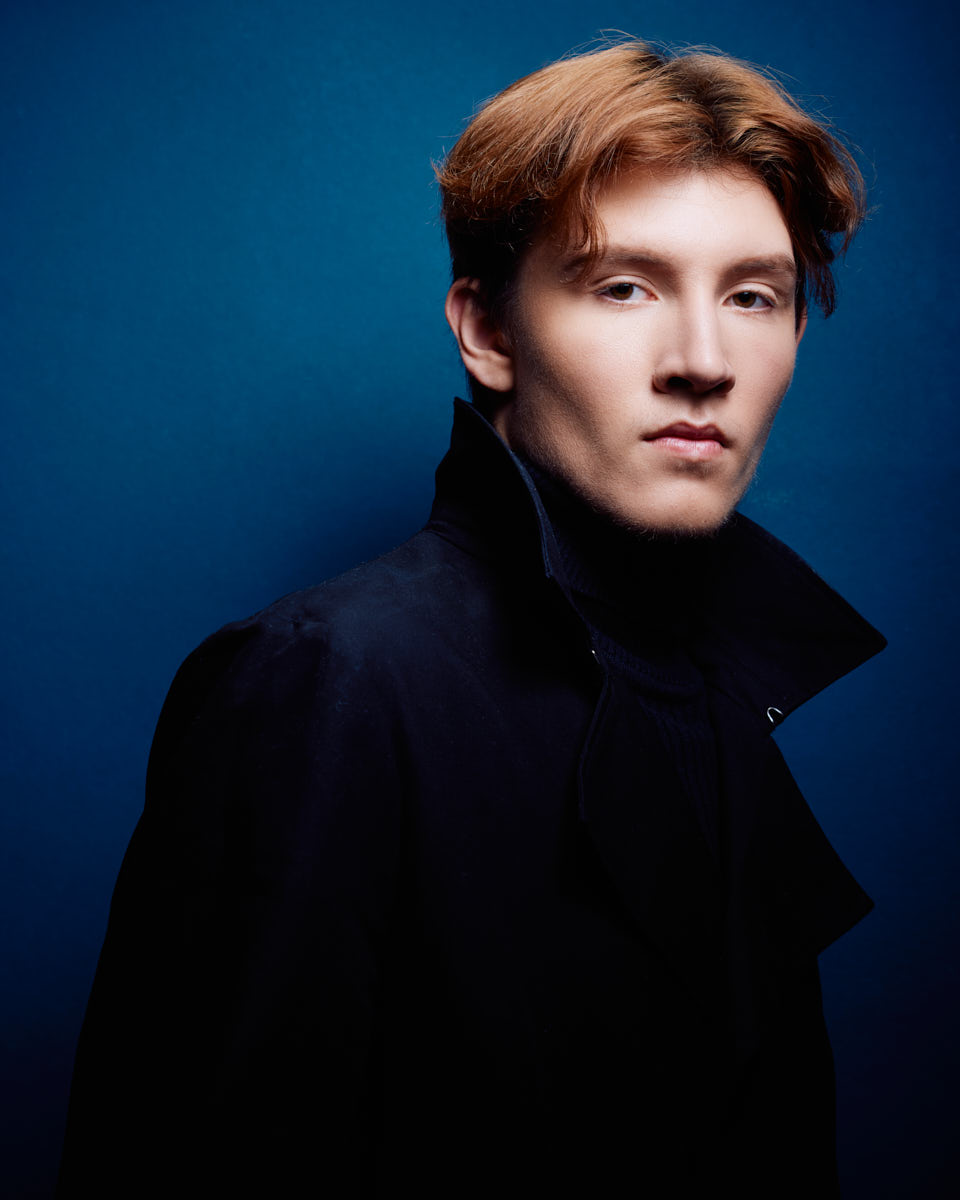 Actor headshots in Chicago featuring Dylan looking serious and stylish in a dark jacket and turtleneck against a dark blue background.