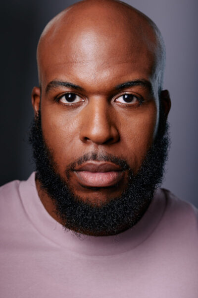 A close-up actors headshots in Chicago of Michael showcasing his intense gaze. He is wearing a light pink t-shirt, and the background is softly lit, focusing all attention on his face. This headshot emphasizes his strong facial features and expressive eyes.