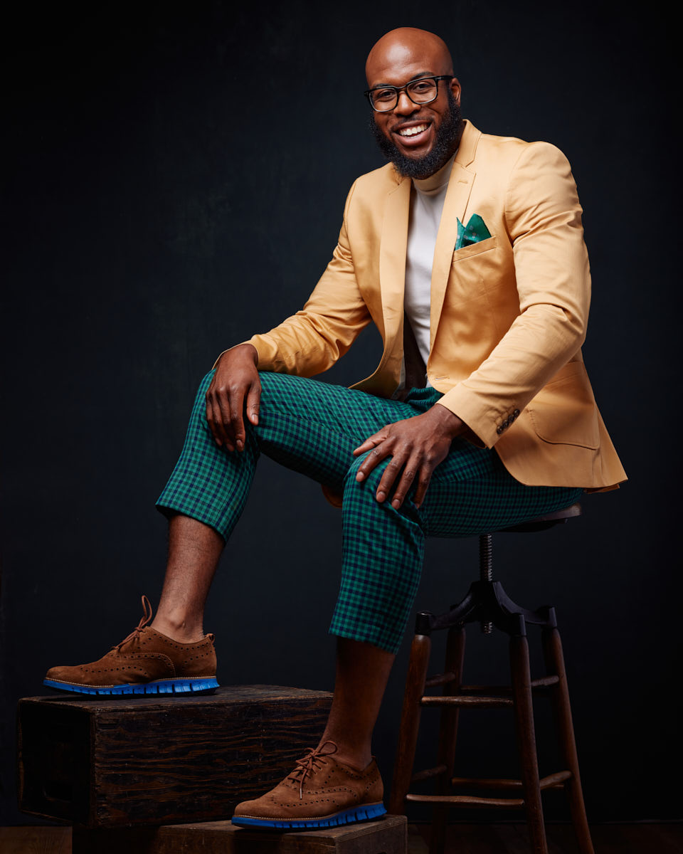 Michael poses in a stylish outfit, consisting of a yellow blazer, green checkered pants, and brown shoes. He is seated on a stool, smiling confidently. The dark background and vibrant outfit make this a standout fashion portrait.
