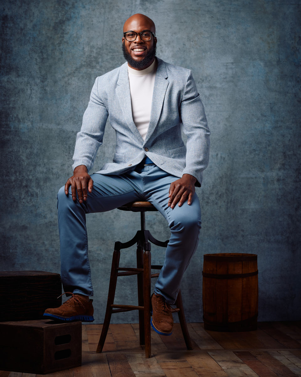 Michael is seated on a stool, wearing a light blue suit with a white t-shirt underneath. He has a relaxed and confident smile, and the setting includes wooden props that add a touch of rustic charm. His outfit and demeanor convey a casual yet professional look.