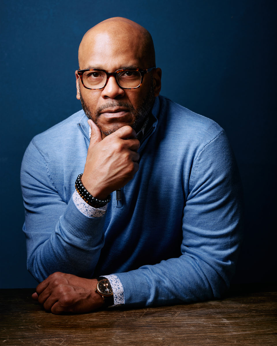 Michael poses with a thoughtful expression, resting his chin on his hand. He is dressed in a light blue sweater, adding a more casual and approachable element to his branding images. The background is a solid dark blue, keeping the focus on him.