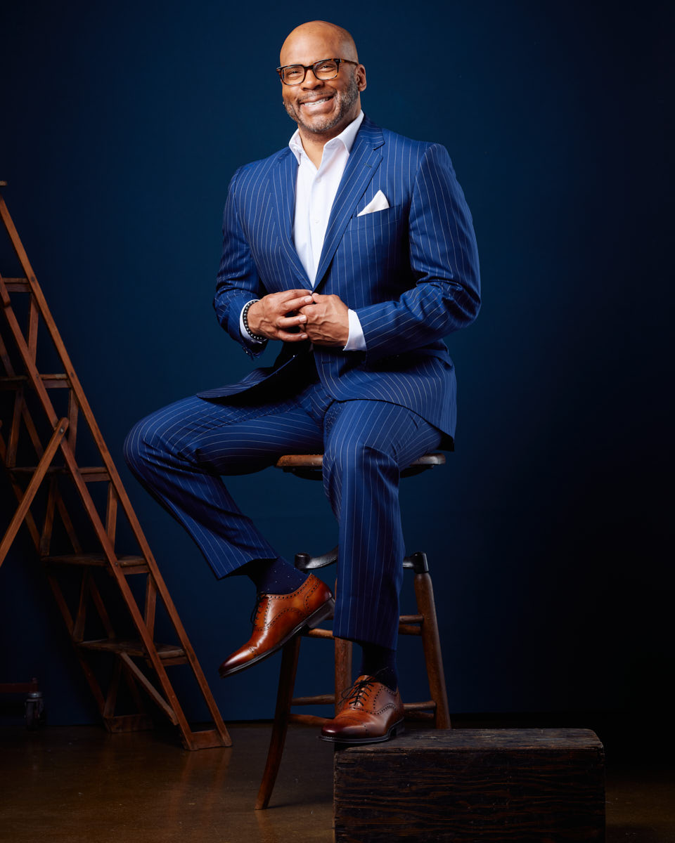 Michael is dressed in a blue pinstripe suit, seated against a dark blue background. His bright smile and confident pose make this a powerful branding image. The contrast between the suit and the background enhances his professional appearance.