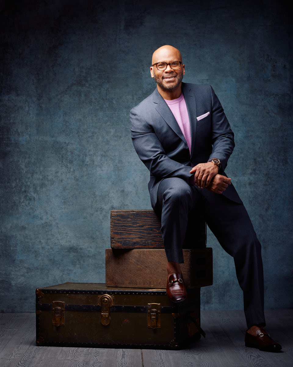 In this image, Michael is seated on a set of wooden boxes, still dressed in his dark suit and light purple sweater. His posture is relaxed yet professional, conveying approachability and confidence. The background remains the same textured gray, providing a consistent theme.