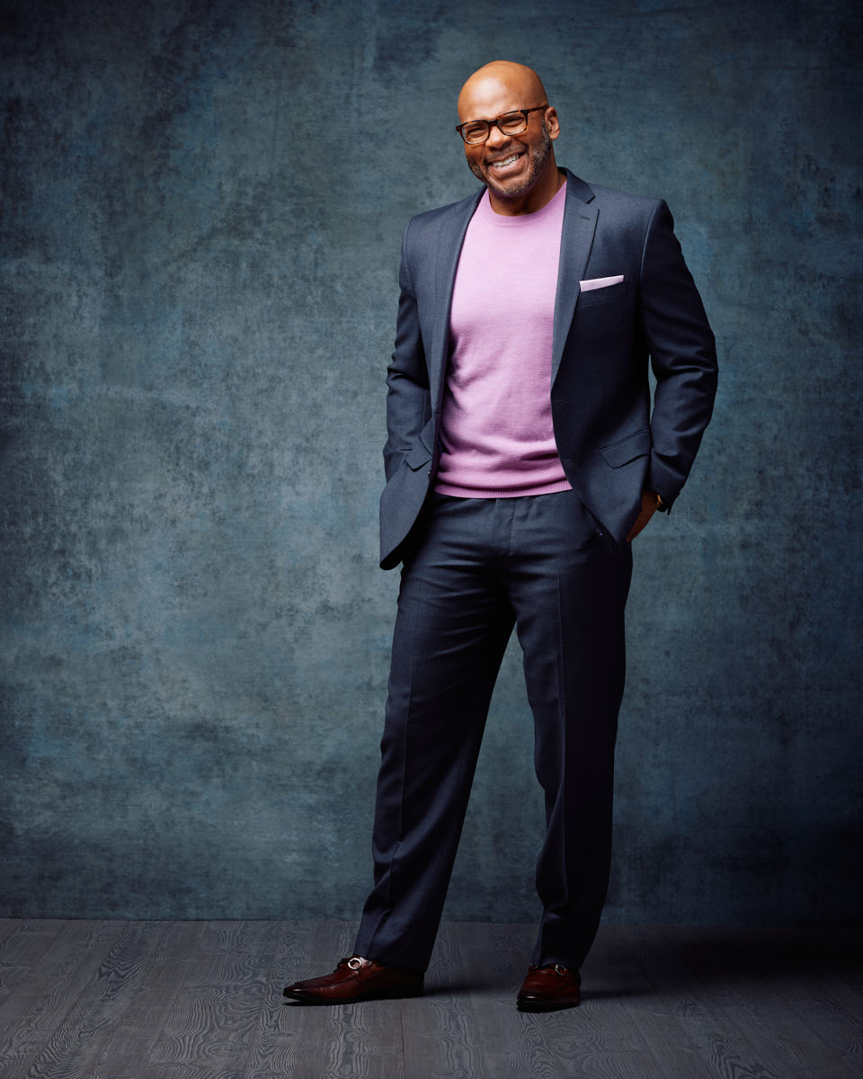 Michael stands confidently against a textured gray background. He is wearing a dark suit paired with a light purple sweater, adding a touch of color to his professional attire. His smile is warm and approachable, ideal for branding and corporate images.
