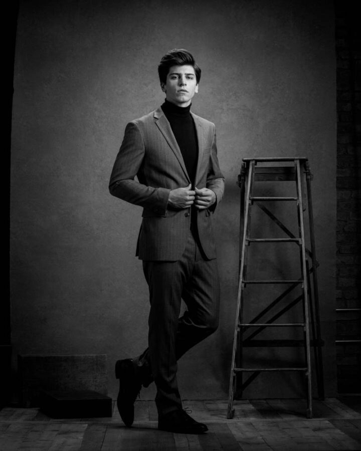 modeling portfolio photographer in Chicago captures Noah is standing in a dark studio setting, wearing a black turtleneck and a gray suit. His pose and the dramatic lighting create a classic and powerful portrait.