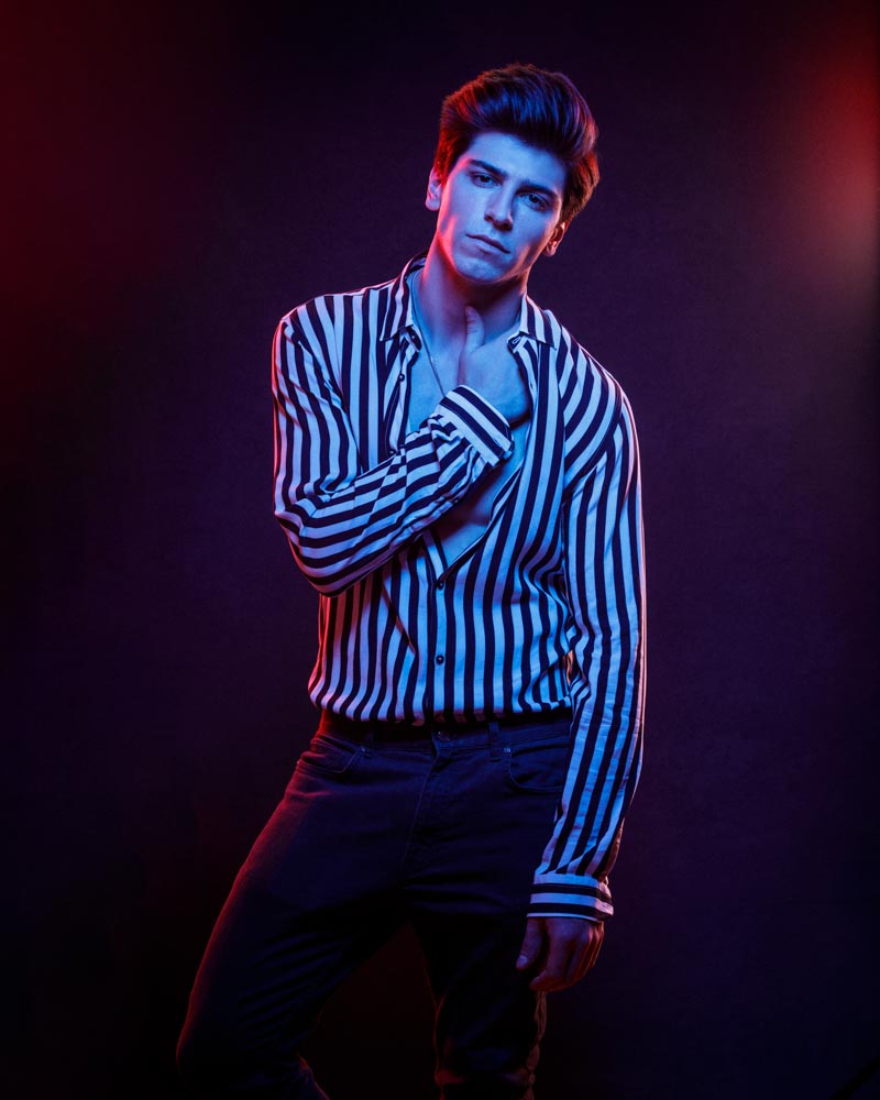 modeling portfolio photographer in Chicago captures Noah is in a blue and black striped shirt, bathed in red and blue lighting. His pose is casual, with one hand on his neck, capturing a modern and edgy look.