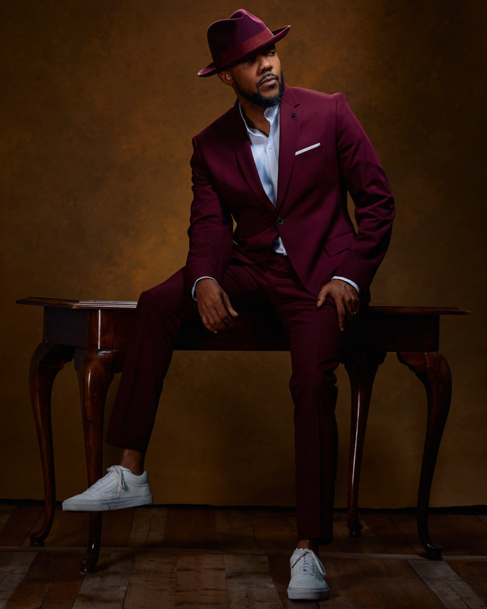 David is sitting on an elegant wooden table, wearing a maroon suit and hat. The rich, warm tones in the background complement his stylish outfit, making this a striking example of male model lookbook photography.