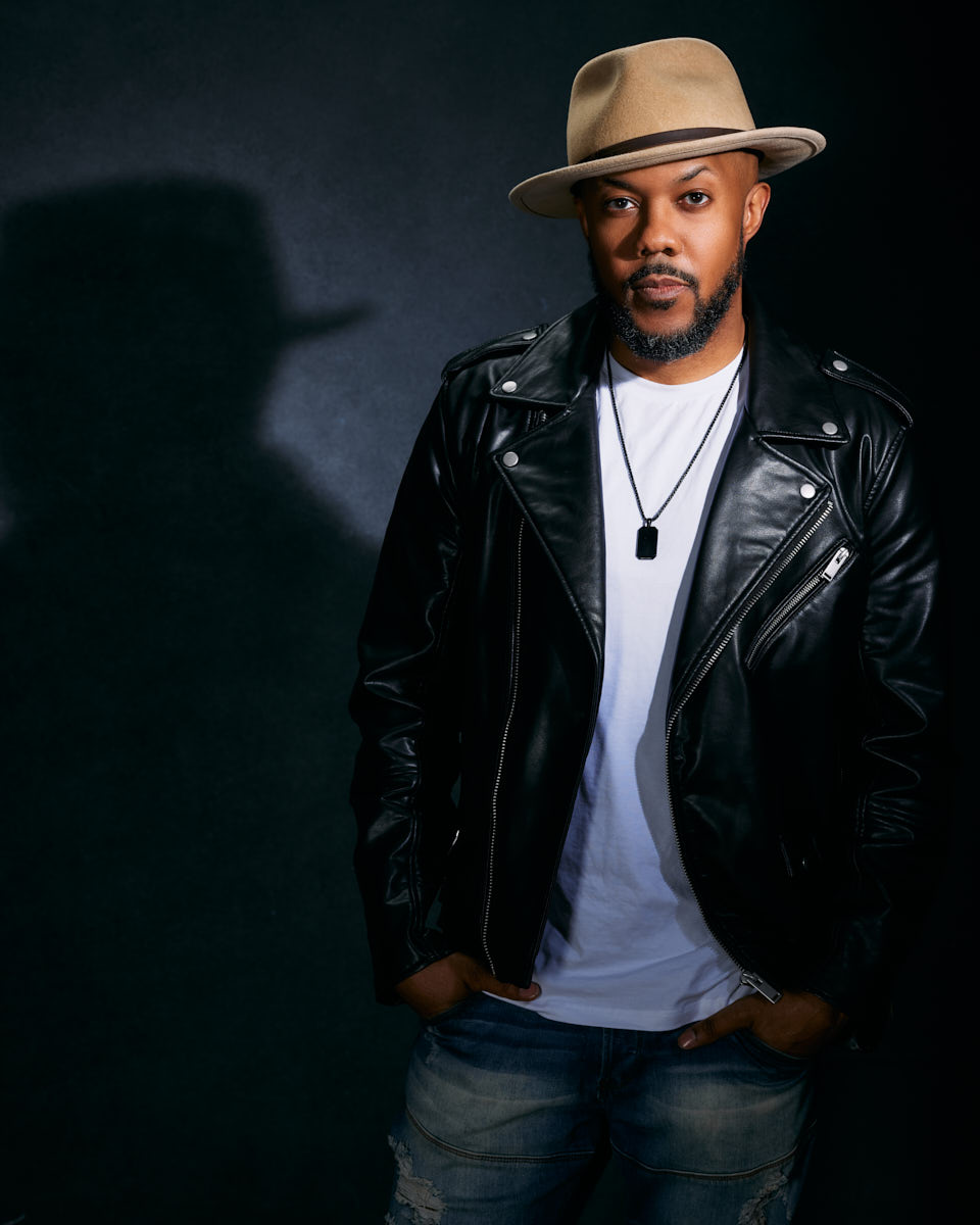 David, in a beige hat and black leather jacket, stands against a dark background. The shadow cast behind him adds a dramatic flair to his overall look.