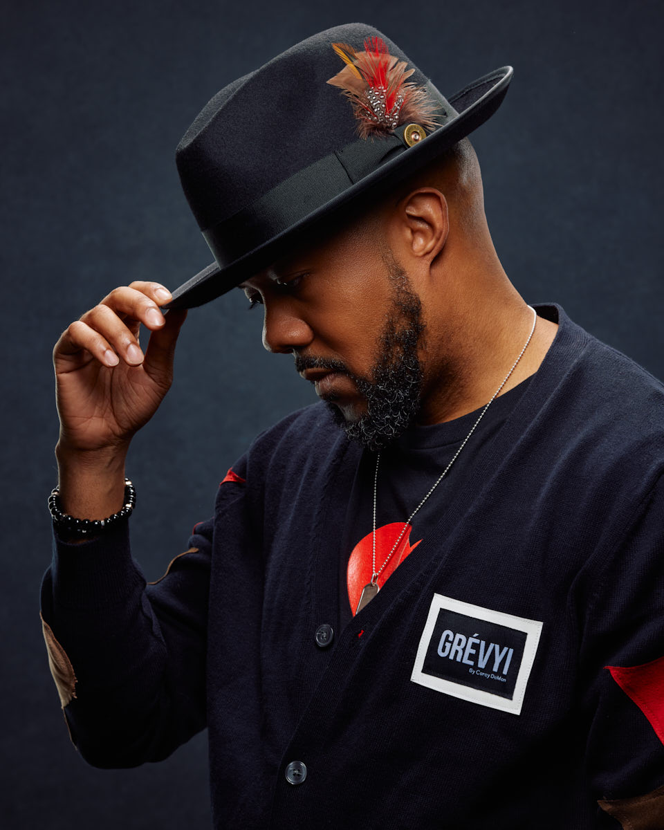 David tips his hat in a profile shot, wearing a black cardigan with a logo patch. This side view, combined with the black background, adds a mysterious element.