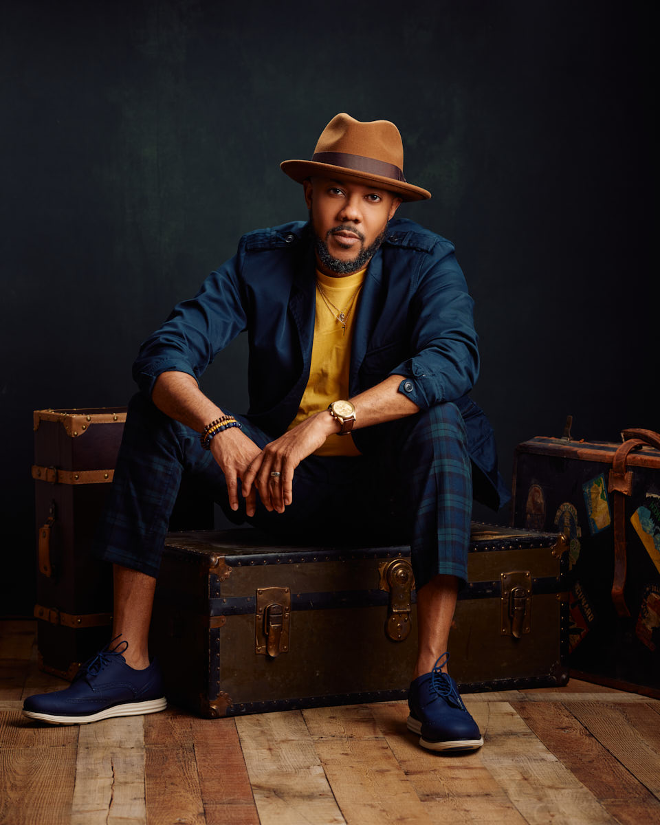 Seated on vintage trunks, David pairs a brown hat with a navy outfit, exuding a stylish, casual look. The earthy tones and relaxed pose showcase his versatility.