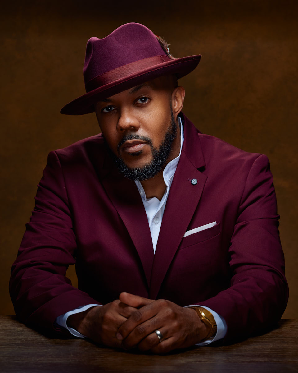 David, dressed in a maroon suit and hat, leans forward with his hands clasped. The warm brown background and his serious expression create a captivating image for male model lookbook photography.