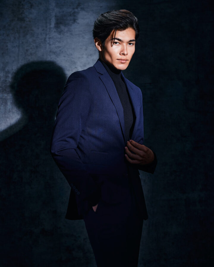 male model portfolio ideas in Chicago featuring Santiago, dressed in a dark blue suit with a black turtleneck, posed against a dark, textured background, creating a dramatic and sophisticated look.