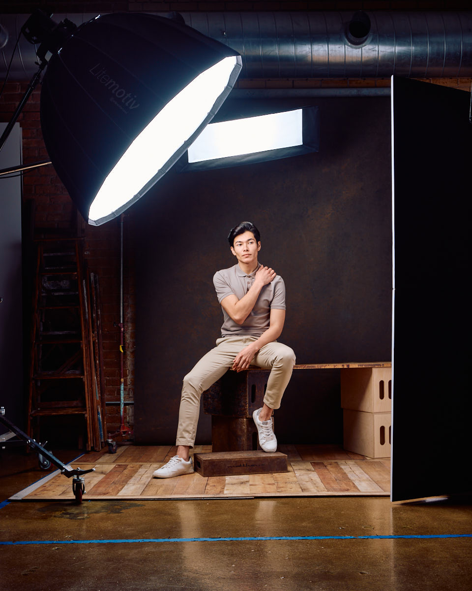 Behind-the-scenes shot of Santiago seated in a studio setting, wearing a grey polo shirt and beige pants. The visible lighting equipment and studio setup offer a glimpse into the professional photoshoot environment.