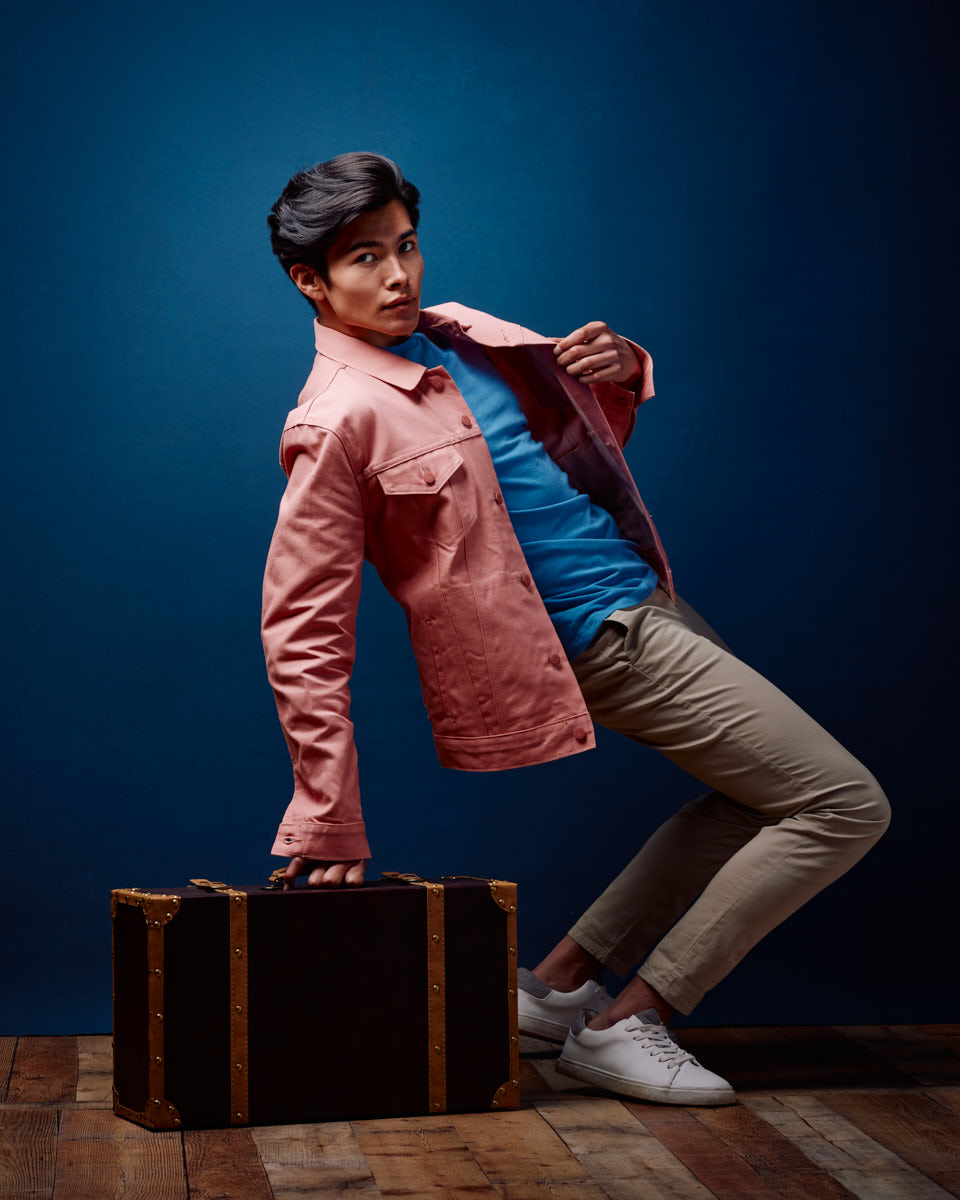 casual lifestyle shots are a great male model portfolio ideas in Chicago. In this image Santiago strikes a dynamic pose, leaning back slightly while holding a vintage suitcase, dressed in a pink jacket, blue shirt, and beige pants. The rustic touch of the wooden floor enhances the scene.