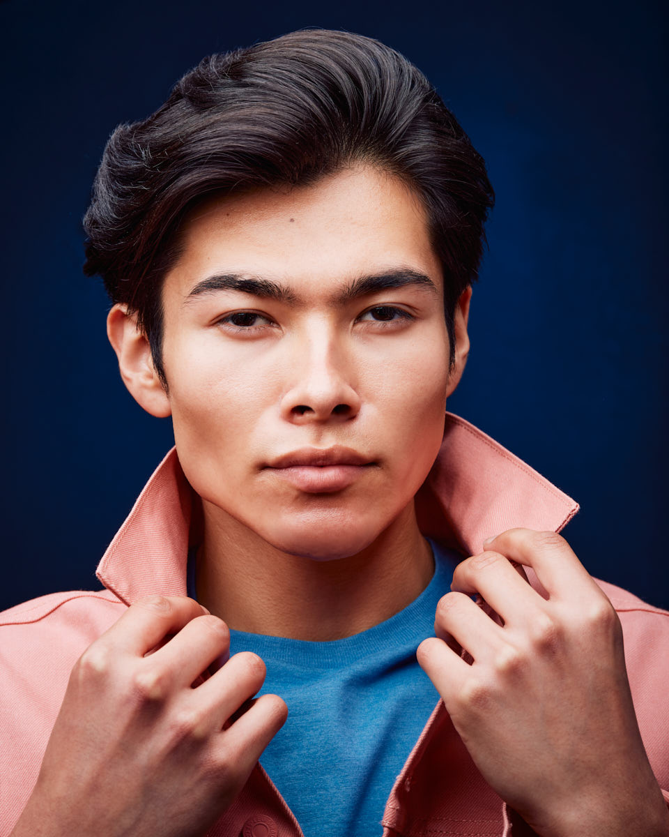 Close-up shot of Santiago in a pink jacket over a blue shirt, with his collar popped. The deep blue background contrasts nicely with his attire, highlighting his facial features and expression.