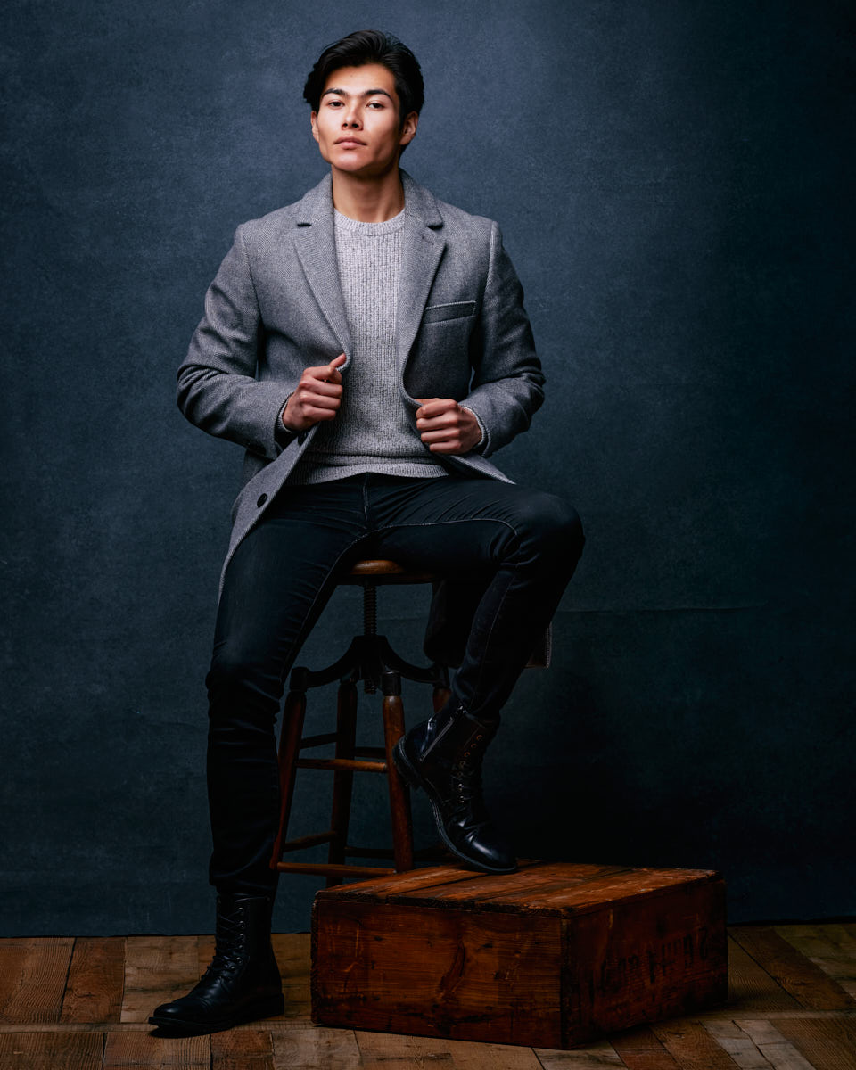 Santiago showcasing his sophisticated look in a gray coat and light gray sweater. The dark, textured background and confident pose emphasize a high-fashion, editorial style.