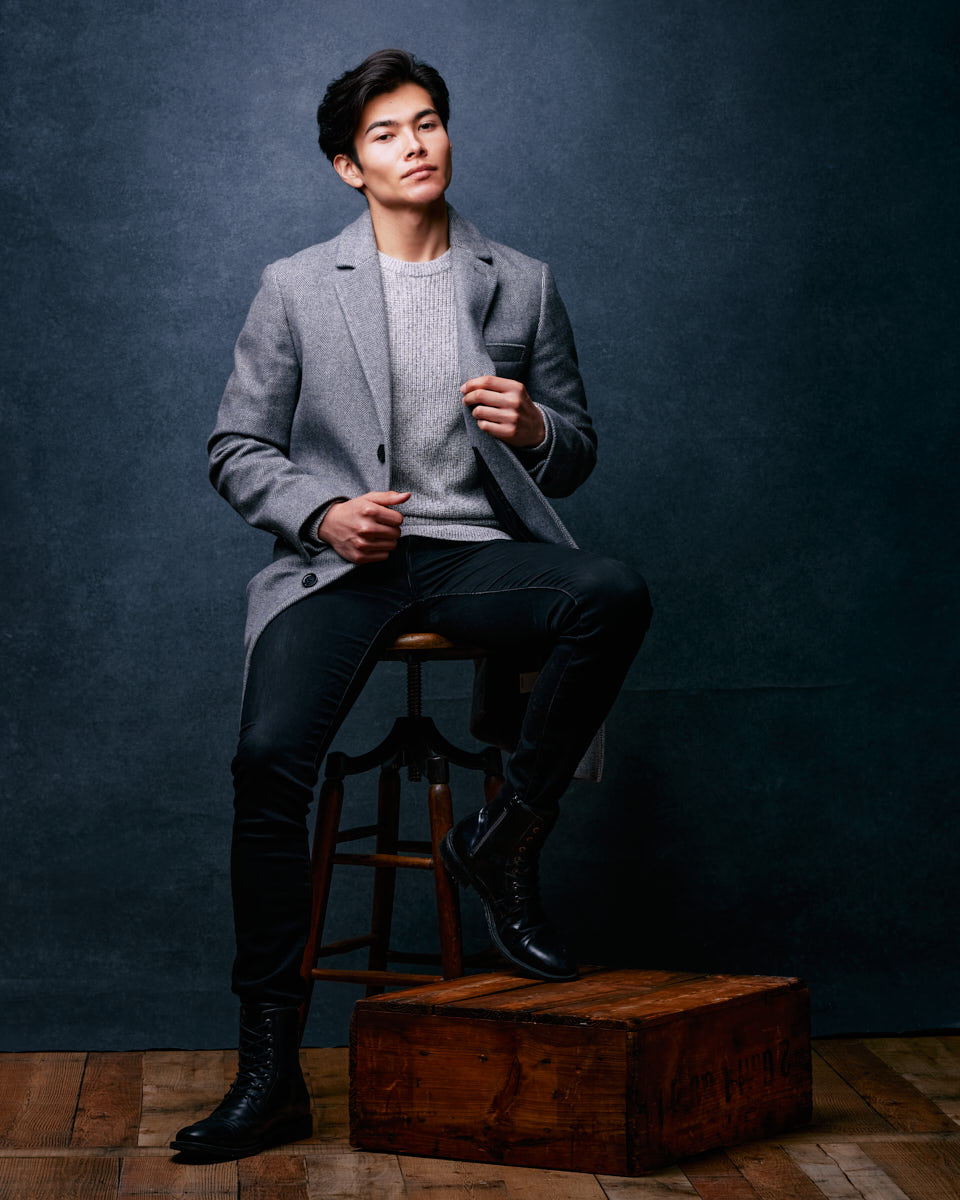 male model portfolio ideas in Chicago featuring Santiago sitting on a stool in a studio environment, wearing a gray coat, light gray sweater, and black jeans. The setup includes professional lighting equipment, adding depth and shadow to the image, perfect for a classic and timeless portfolio shot.