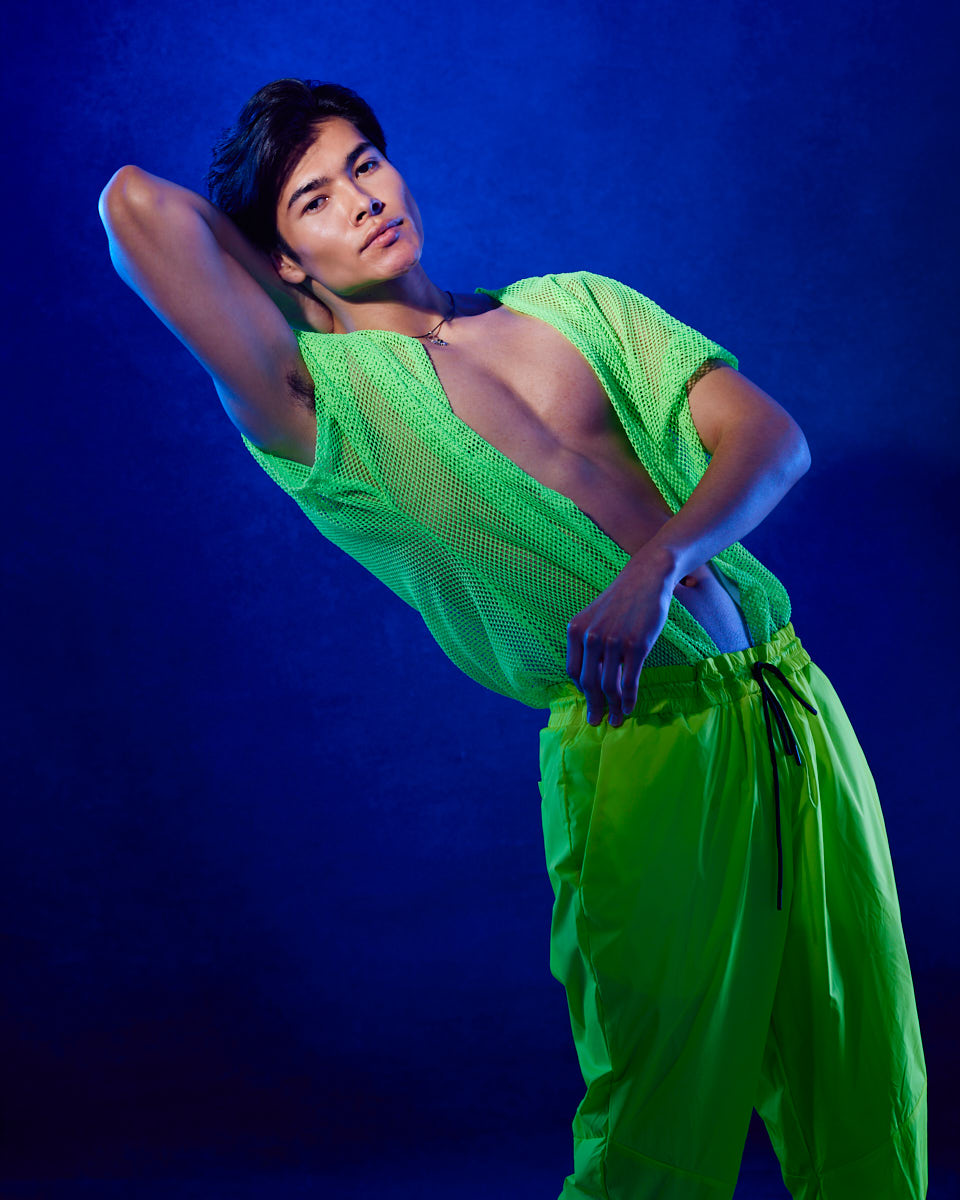 male model portfolio ideas in Chicago featuring Santiago strikes a bold and confident pose, wearing a neon green mesh top that is partially open, revealing his torso. The background is a rich blue, making the neon green outfit pop. The lighting highlights his defined features and athletic build, showcasing versatility in high-fashion modeling.