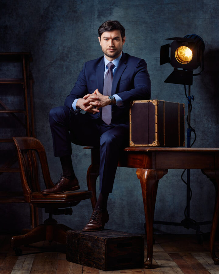 Stanislav is seated at a vintage wooden table, dressed in a sharp navy suit with a tie. The setting includes a classic spotlight and antique trunks, adding a touch of old-world charm to the modern, professional portrait for his male model portfolio in Chicago