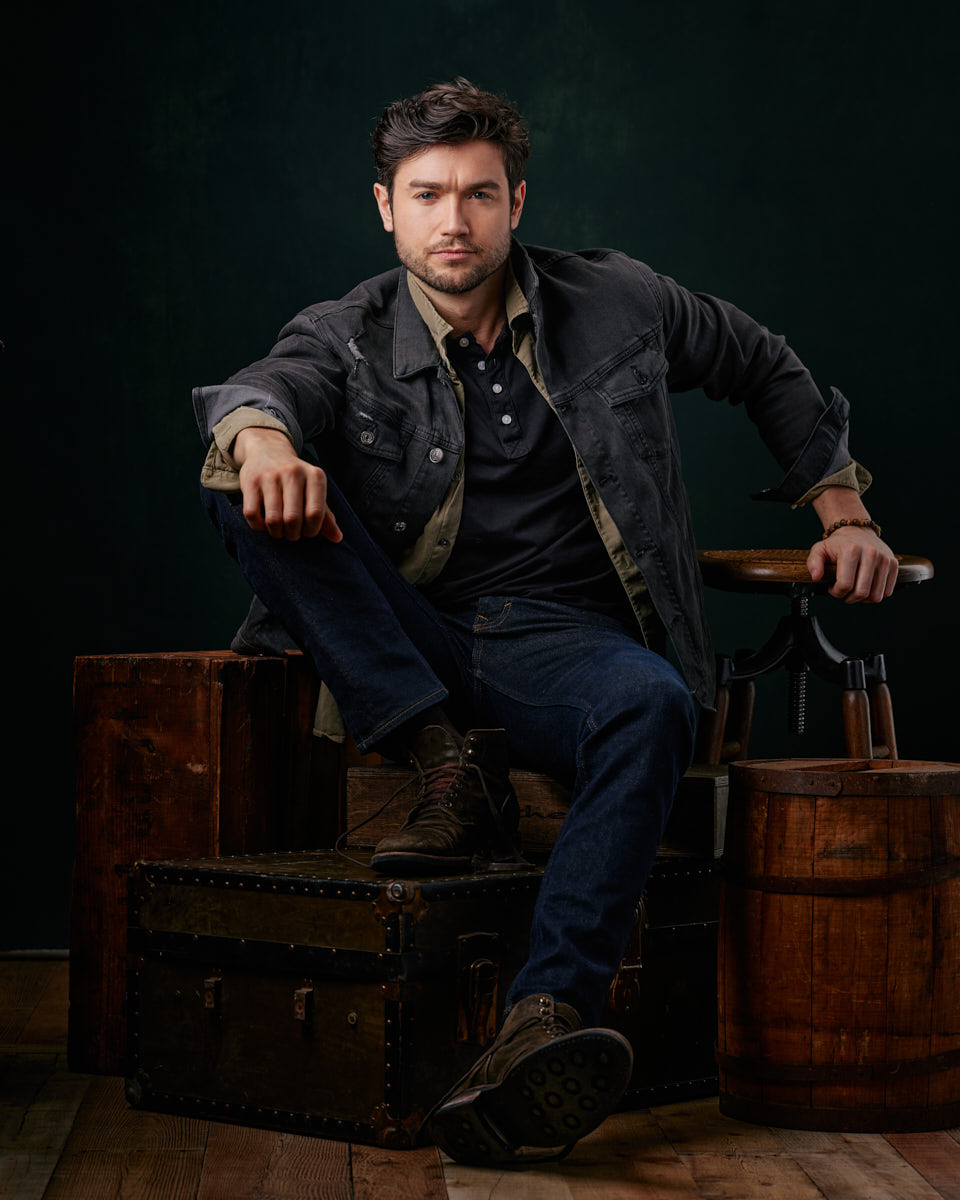 Stanislav is in a rugged, casual look, sitting on stacked vintage trunks. He wears a dark jacket over a henley shirt and jeans, giving off a rugged and approachable vibe. His pose is relaxed, hands clasped, and he looks confidently into the camera. male model portfolio in Chicago