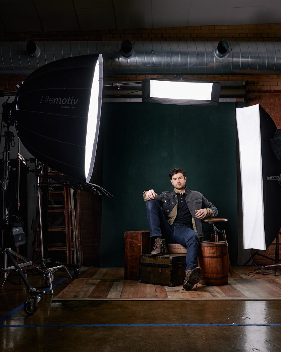 Stanislav sitting casually on stacked wooden crates in a studio setup. He is wearing a denim jacket over a dark shirt and jeans, exuding a rugged, stylish look. The studio lighting and equipment are visible, emphasizing the professional setting