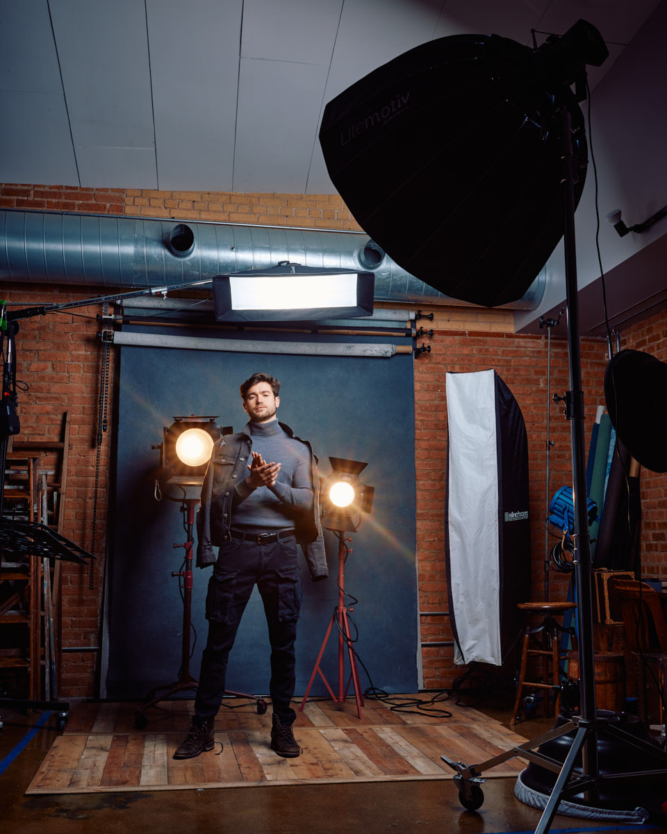 Stanislav is fully dressed in a gray turtleneck and black cargo pants, standing in front of two vintage studio lights. The lighting setup is visible, creating a dramatic effect with the warm glow of the lights