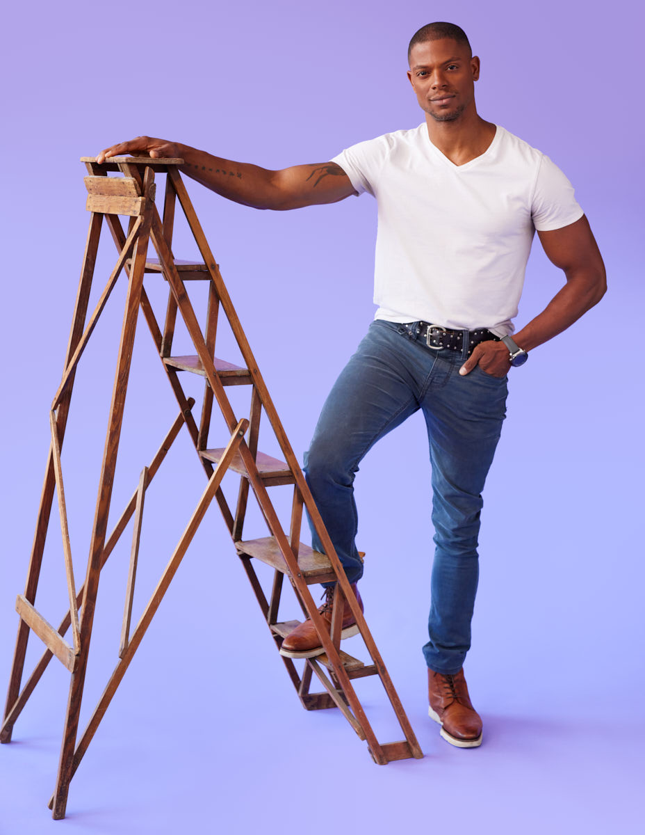 Jason is standing with one foot on a wooden ladder against a light purple background.