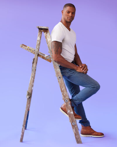 male model posing guide in Chicago featuring Jason leaning against a wooden ladder, with his back resting on the side and his left leg bent, foot resting on the bottom rung. He is wearing a white t-shirt, blue jeans, and brown leather boots. His hands are clasped in front of him, and he is looking slightly to the side. Jason’s pose is casual and comfortable, with a hint of a smile on his face.