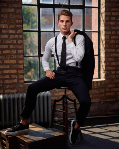 Christian standing in a gray suit with a burgundy tie. He leans casually against a chair with one hand in his pocket. The setup includes professional studio lighting and a backdrop, with large windows providing additional light which meets the male model requirements in Chicago