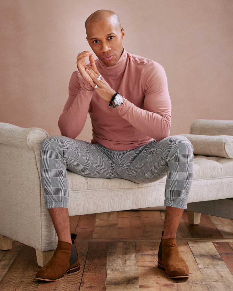 A color photo of the male model sitting on a light-colored couch. He is dressed in a pink turtleneck and checkered pants with brown boots. He has a watch and bracelets on his left wrist, and he is looking directly at the camera with his hands clasped.
