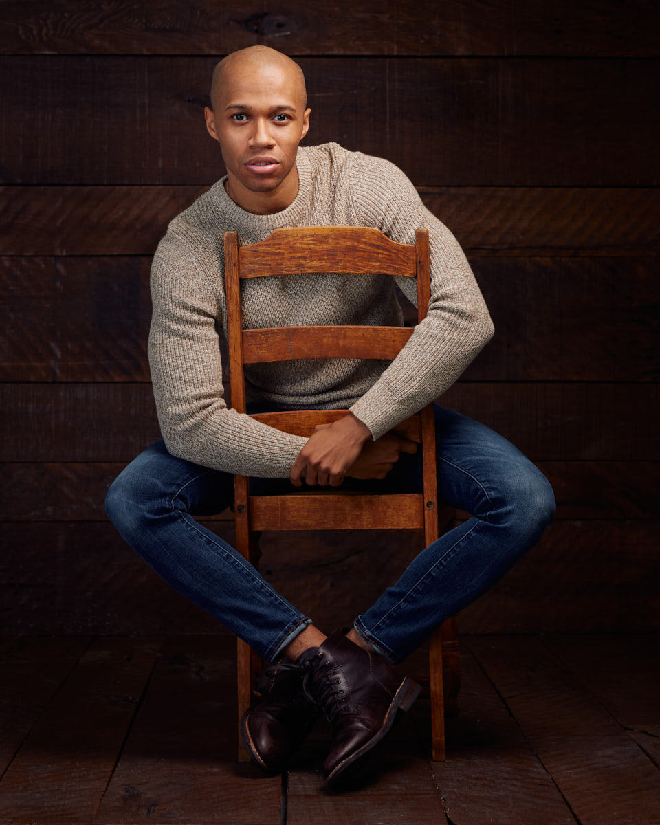 A color photo of the male model sitting on a wooden chair against a wooden background. He is wearing a beige sweater and blue jeans, looking directly at the camera with a relaxed and approachable expression.