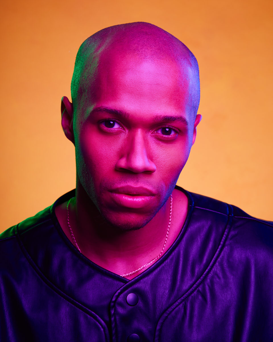 A close-up color photo of the male model against a yellow background. He is wearing a leather jacket and the lighting is a mix of pink and green, giving a dramatic effect. He is looking directly at the camera with a serious expression.