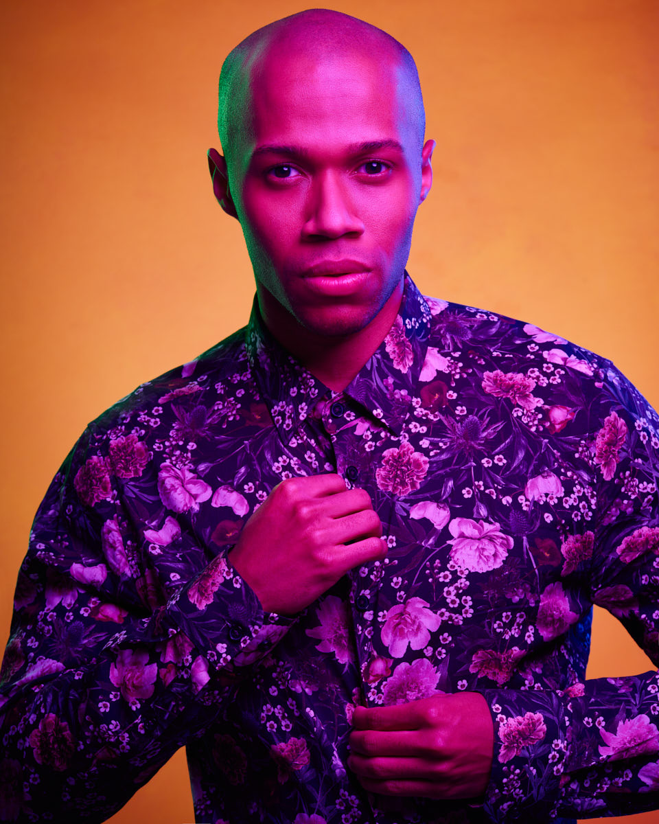 A vibrant color photo of the male model in a floral shirt with a yellow background. The lighting is a mix of pink and green, giving the image a dramatic and artistic feel. He is buttoning his shirt and looking directly at the camera.