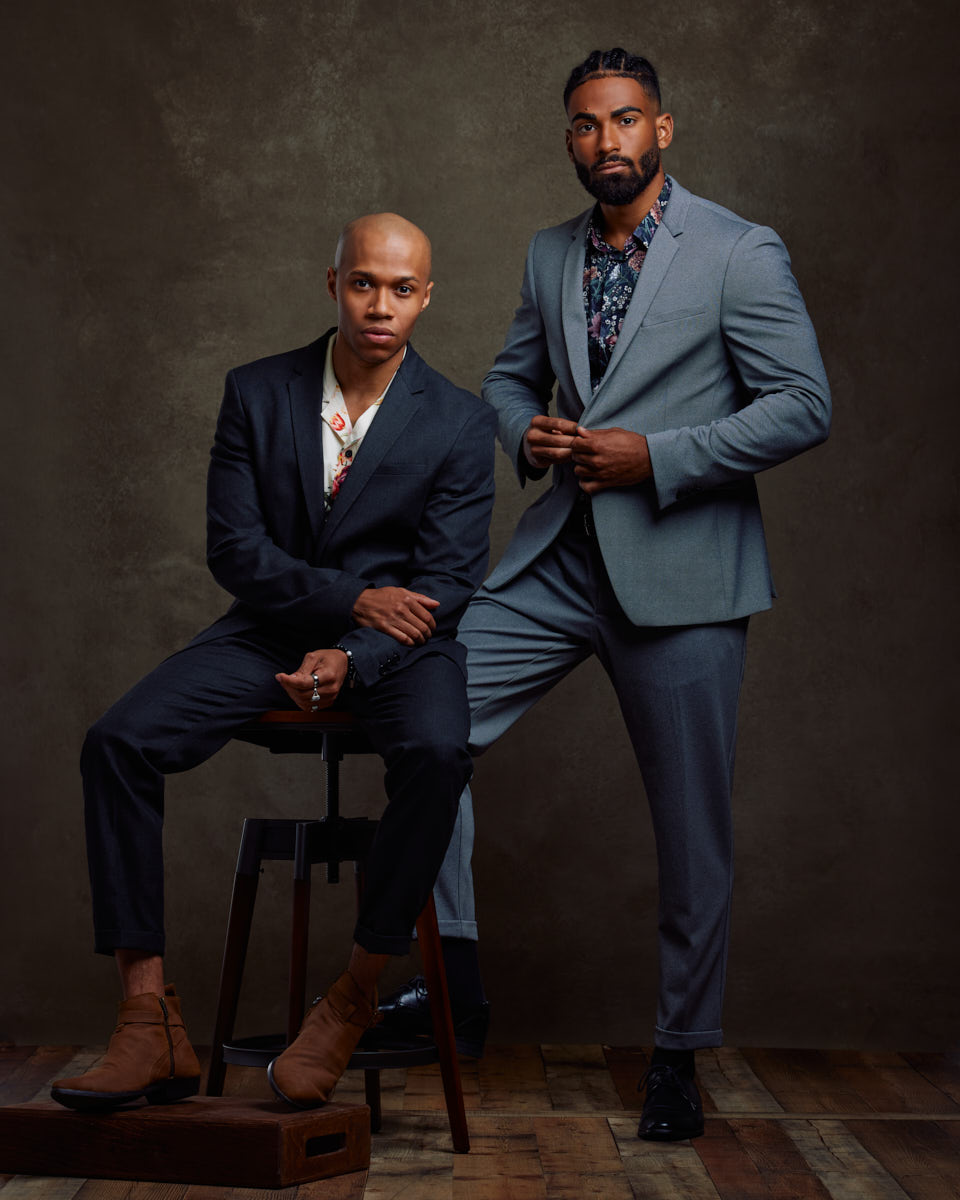 A color photo of two male models in suits. The male model from the previous images is seated on a stool while another model stands beside him. Both are dressed in suits with patterned shirts and brown shoes, posing confidently against a dark backdrop.