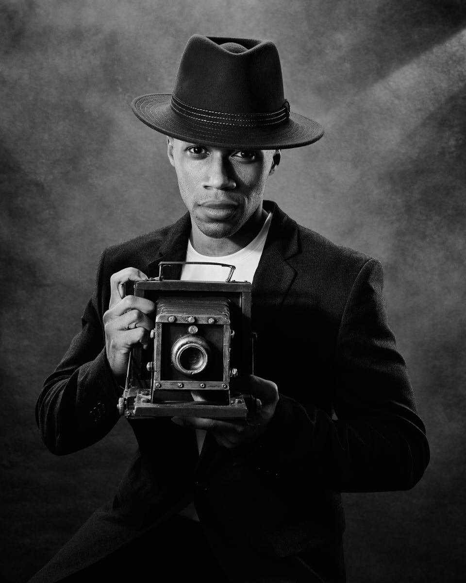 A black-and-white close-up of the male model holding a vintage camera. He is wearing a suit and a hat, looking directly at the camera with a serious expression.