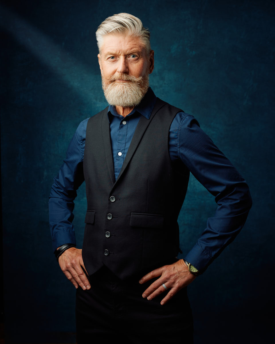 A mature male model with a beard stands confidently with his hands on his hips. He is wearing a dark blue shirt and a black vest, with a subtle smile and a piercing gaze. The background is a rich, dark blue, adding depth to the portrait.