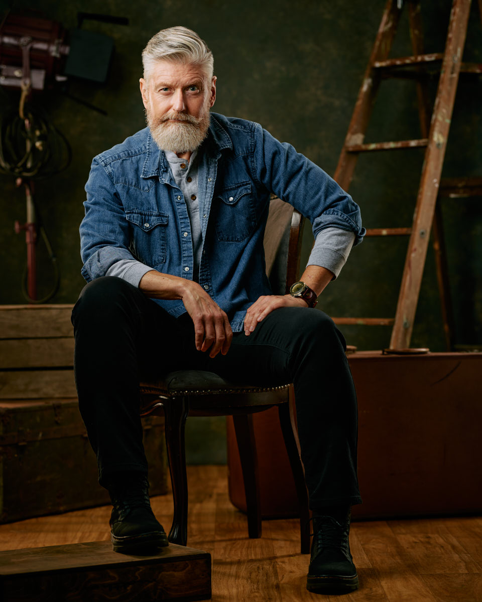 Jeff poses confidently on a chair, wearing a denim shirt and dark pants, with a serious expression and vintage props in the background a great image for the male modeling industry in Chicago