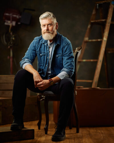 Full body portraits are standard in the male modeling industry in Chicago. Jeff sits on a chair in a rustic studio setting, dressed in a denim shirt, dark pants, and black boots, with a ladder and vintage equipment in the background.
