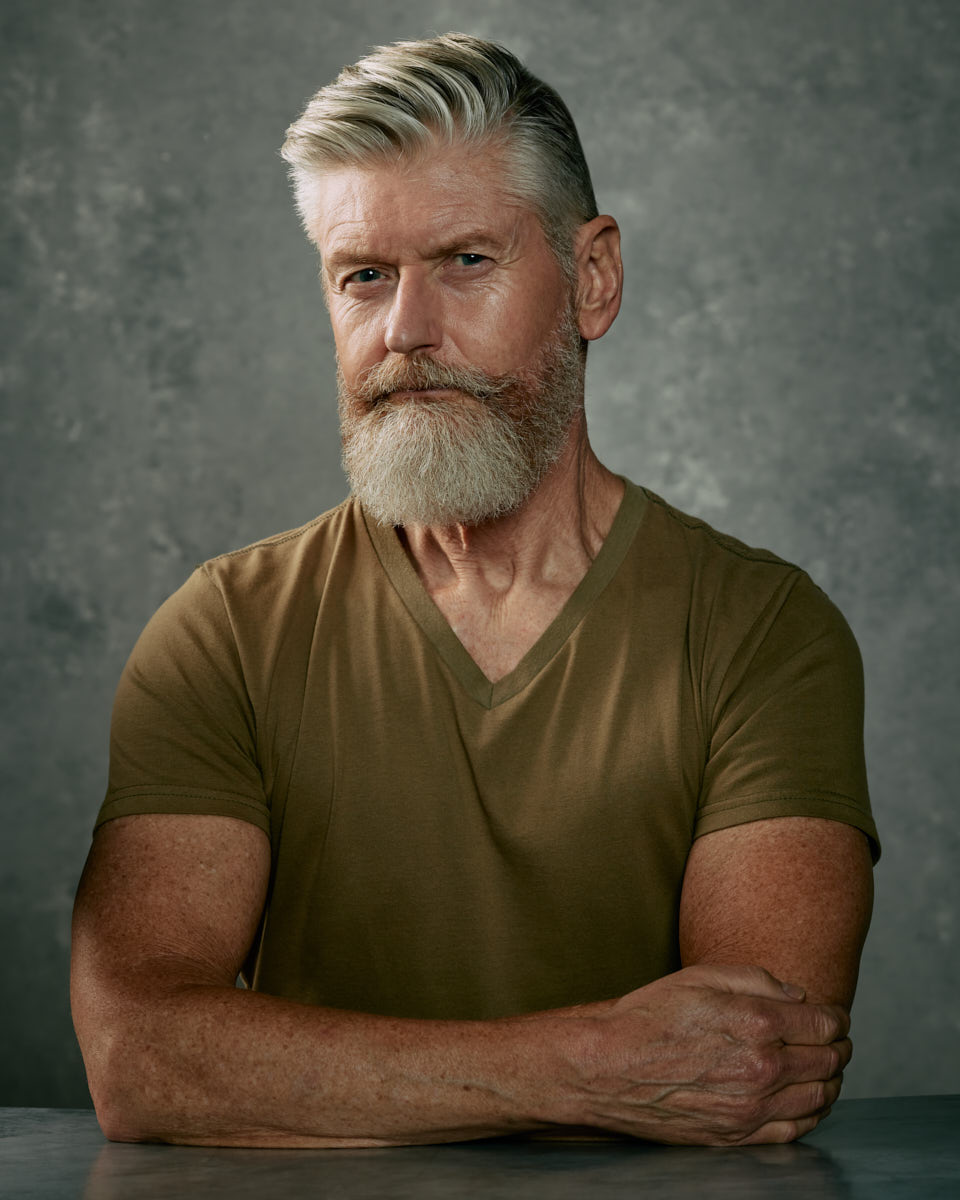 Waist up portraits are standard in the male modeling industry in Chicago. Jeff poses with his arms crossed, wearing a green V-neck shirt, against a simple grey background, highlighting his strong features and well-groomed beard.