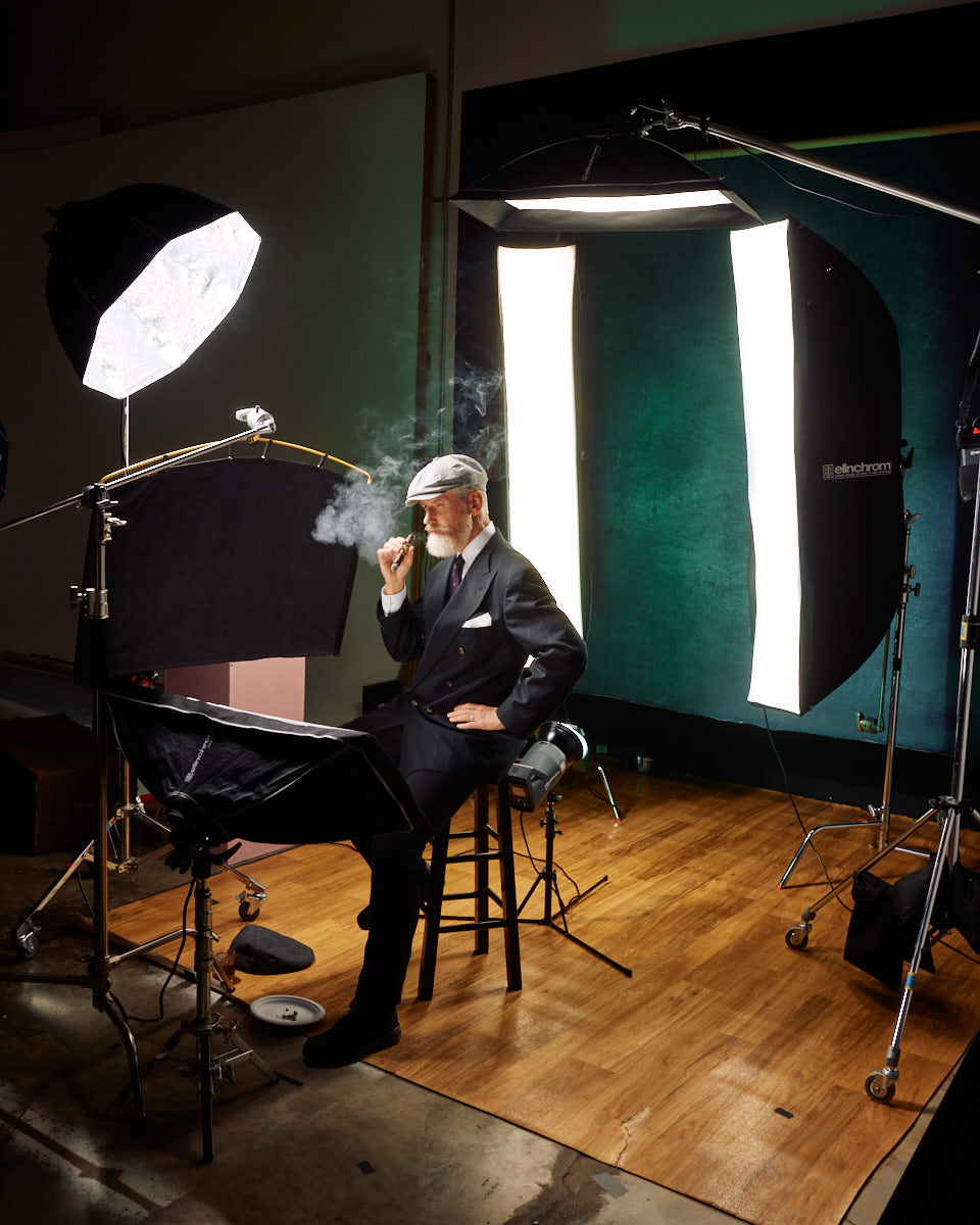 A behind-the-scenes shot of the model in a studio setting. He is sitting on a stool, smoking a cigar, with a lighting setup around him. The scene includes large softboxes and other lighting equipment, highlighting the professional environment.