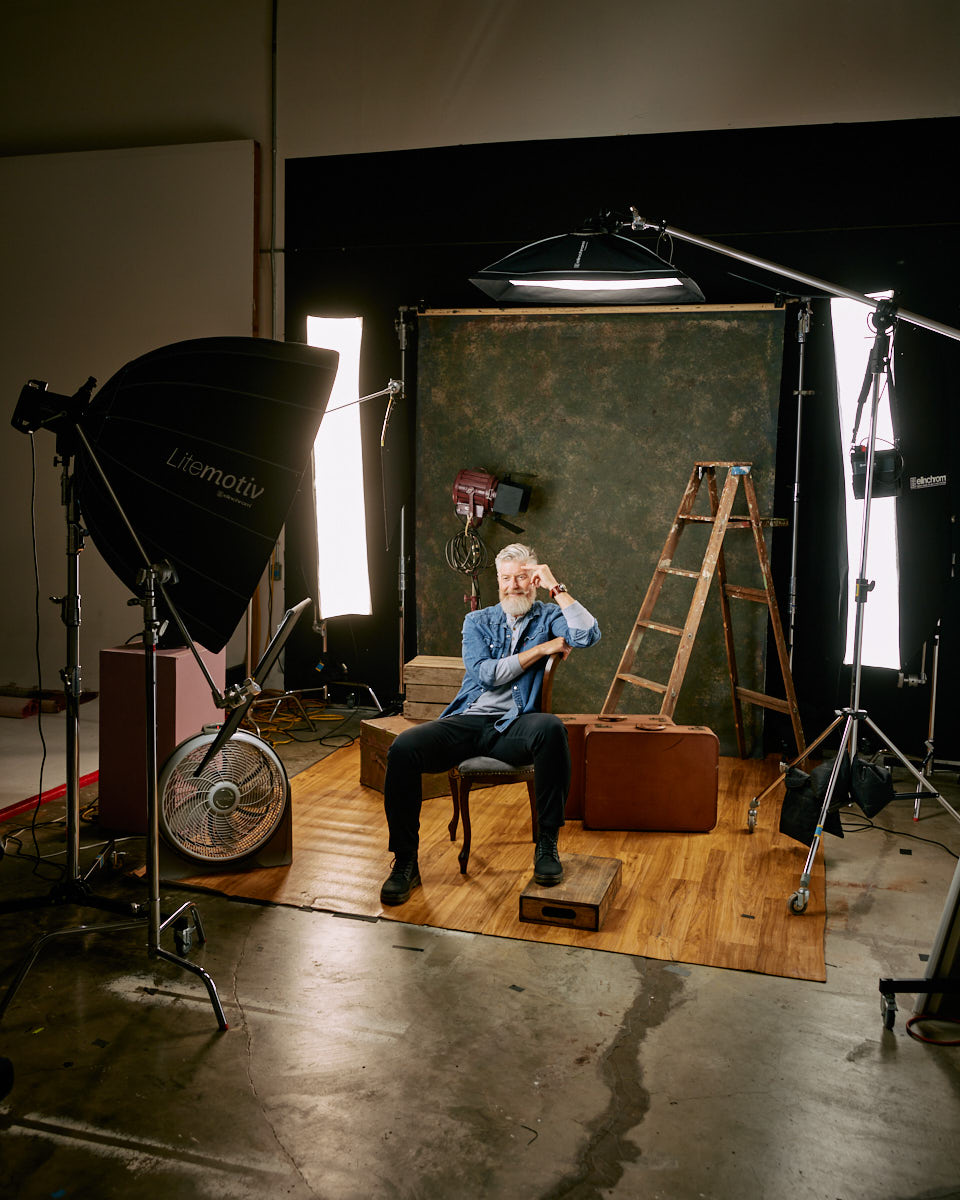 A behind-the-scenes shot of Jeff in the studio, sitting on a chair with a smile, surrounded by professional lighting equipment and a rustic backdrop.