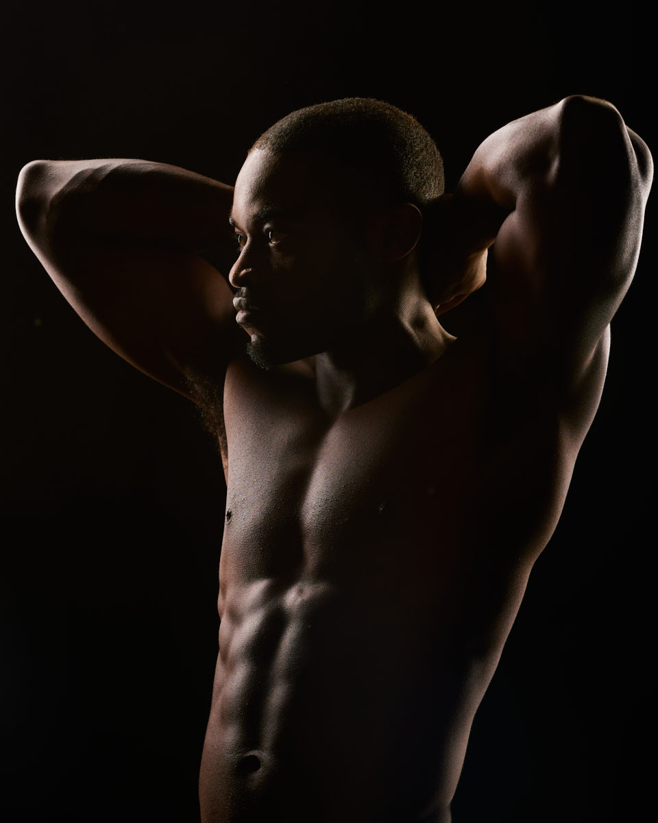 A dramatic, low-light profile shot of Chido, showcasing his muscular physique as he poses with his hands behind his head, emphasizing the contours of his body.